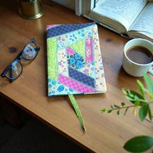 Load image into Gallery viewer, Colorful patchwork notebook on a wooden desk with glasses, a cup of coffee, and an open book.

